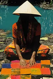 Midjourney generated image using SREF code Polychromaze: A woman in a conical hat sitting on the ground next to a river.