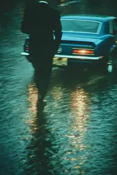 Midjourney generated image using SREF code Blue Velvet: A man in a suit walking down a wet street next to a car.